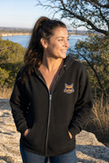 Blind Kitty Zip Hoodie in Black on a woman above Lake Travis, the Blind Kitty on the left chest (AI-generated scene)