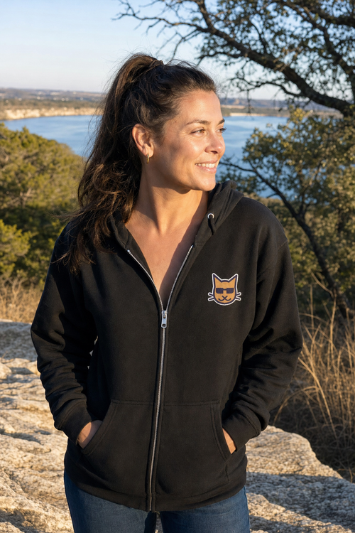 Blind Kitty Zip Hoodie in Black on a woman above Lake Travis, the Blind Kitty on the left chest (AI-generated scene)