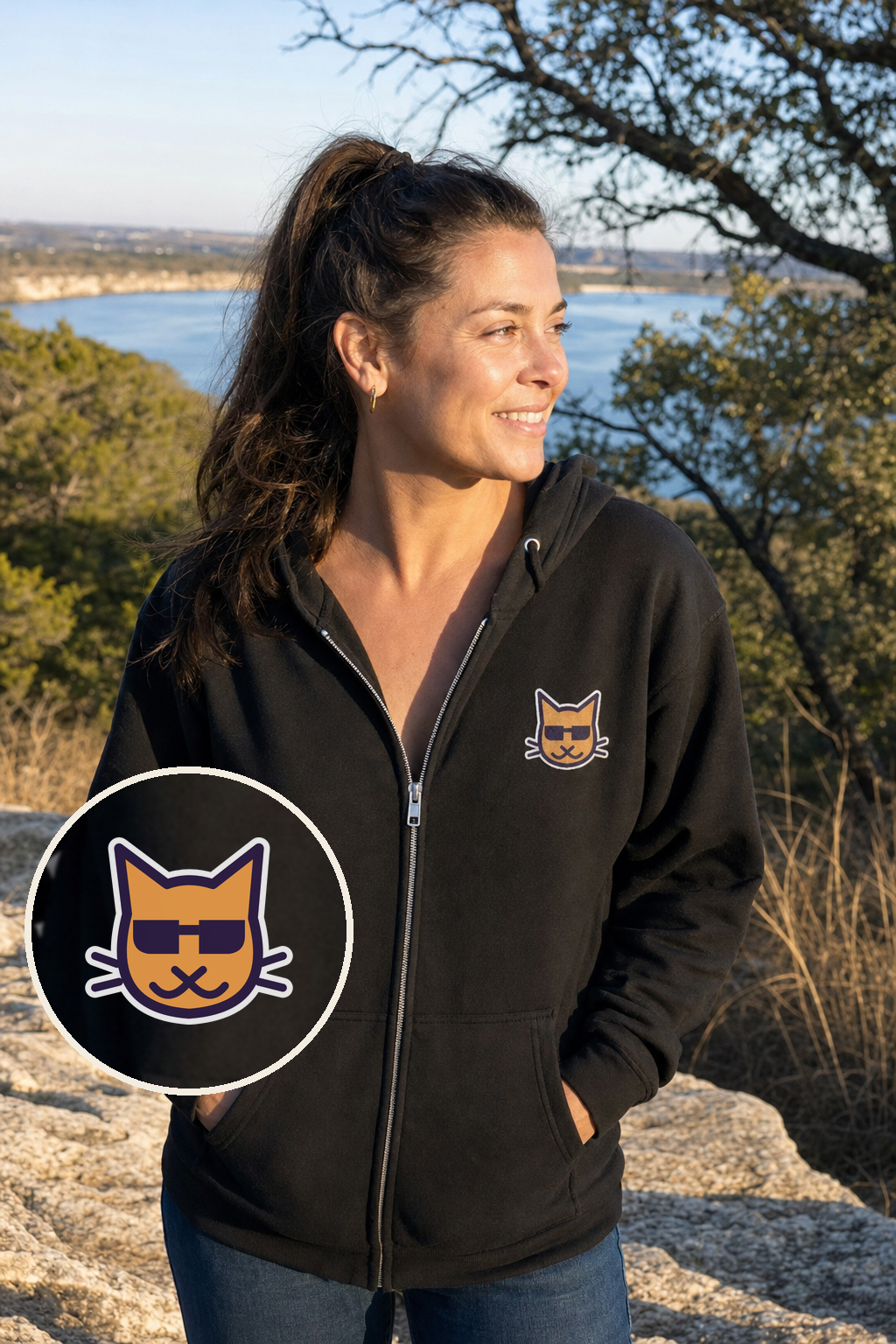 Blind Kitty Zip Hoodie in Black on a woman above Lake Travis, with a magnified close-up of the small Blind Kitty cat printed on the left chest (AI-generated scene)